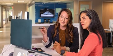 O mentoring e o coaching executivo são processos de desenvolvimento pessoal. Descrição da imagem: duas mulheres de negócios conversando enquanto olham um computador.