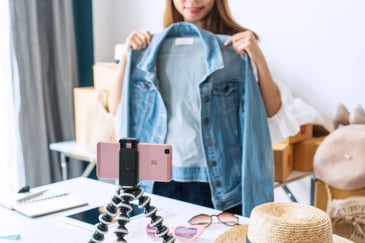 social commerce - jovem segurando jaqueta jeans em frente ao celular que está em um mini tripé