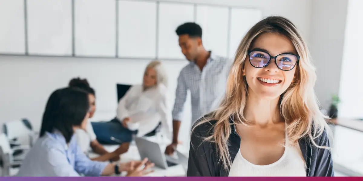 PMO é a sigla para Project Management Office. - Mulher de óculos sorrindo em primeiro plano, em uma sala de reuniões com pessoas.